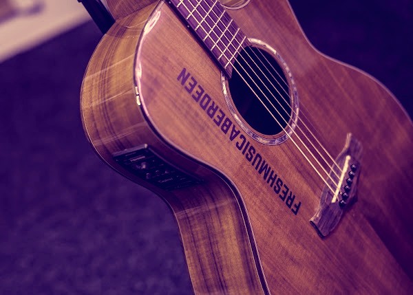 Close-up of a wooden acoustic guitar with FRESHMUSICABERDEEN printed on its body, lit with soft purple light