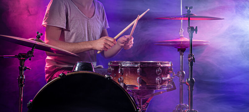 A drummer playing an acoustic drum kit under blue and pink lights with smoke in the background
