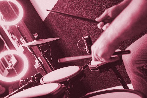 A drummer playing an electronic drum kit with sticks in hand, beside glowing ring lights, in a pink-toned studio shot