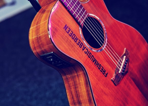 Angled close-up of a glossy orange-toned acoustic guitar with Fresh Music Aberdeen printed across its body