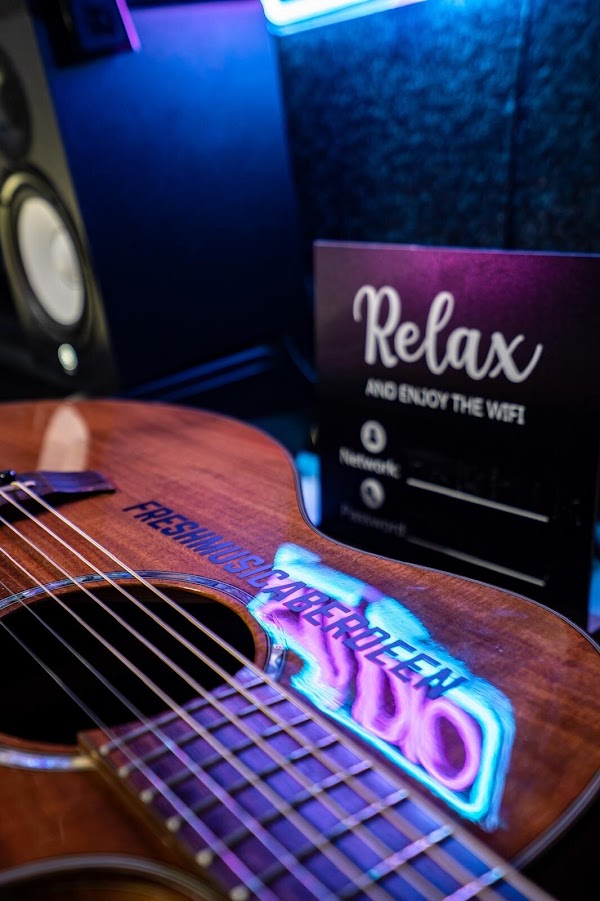 Close-up of an acoustic guitar with Fresh Music Aberdeen printed on its body, reflecting a pink and blue neon Studio sign, beside a studio speaker and a Relax and enjoy the wifi sign