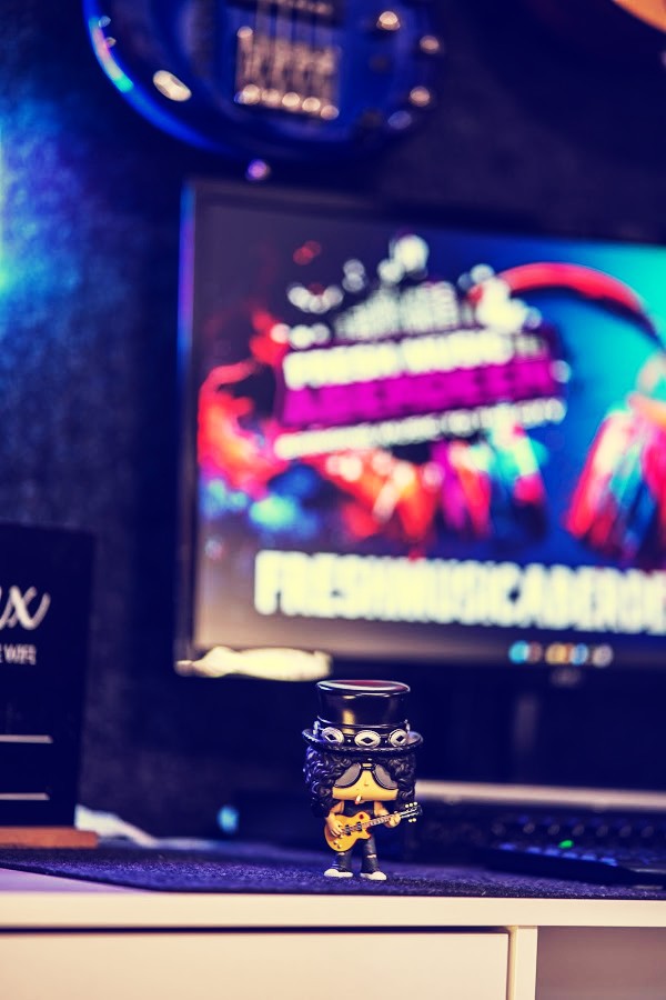 Small collectible figure of a top-hatted guitarist holding a gold guitar on the studio desk, in front of a blurred screen showing the Fresh Music Aberdeen logo