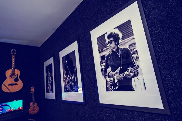 Framed black and white portraits of famous guitarists on the dark acoustic wall of the studio, with an acoustic guitar and a mandolin hanging in the corner