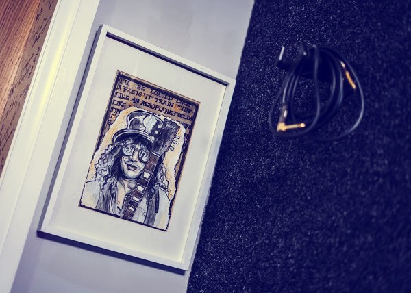 Framed hand-drawn sketch of a curly-haired guitarist in a top hat and sunglasses leaning against the wall, beside a coiled guitar cable on the carpet