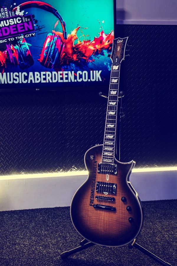 Sunburst LTD electric guitar with Fresh Music Aberdeen printed on the body, on a stand beneath a wall-mounted screen showing the freshmusicaberdeen.co.uk website