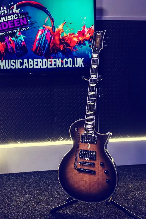 Sunburst LTD electric guitar with Fresh Music Aberdeen printed on the body, on a stand beneath a wall-mounted screen showing the freshmusicaberdeen.co.uk website