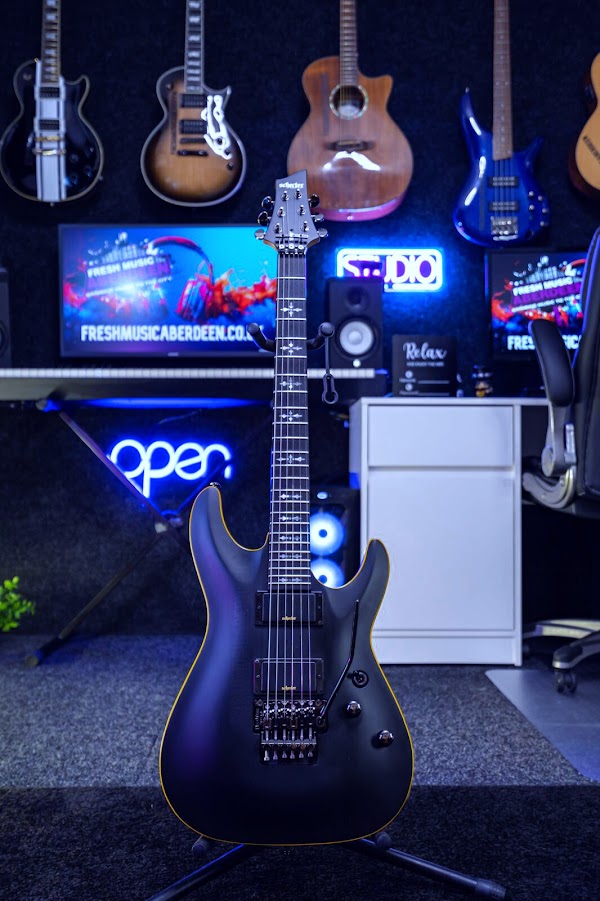 A black Schecter electric guitar on a stand in the Fresh Music Aberdeen studio, with electric, acoustic and bass guitars on the wall behind, a Fresh Music screen, studio monitors and blue neon open and studio signs