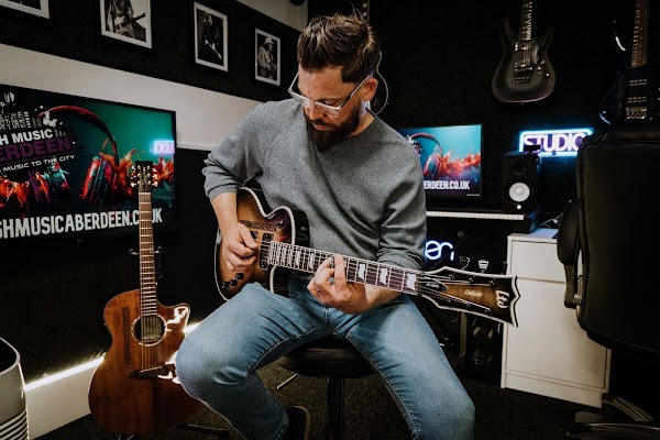 Guitar tutor wearing glasses playing a sunburst electric guitar on a stool in the studio, with an acoustic guitar, framed photos, neon Studio sign and screens showing the Fresh Music Aberdeen logo behind him