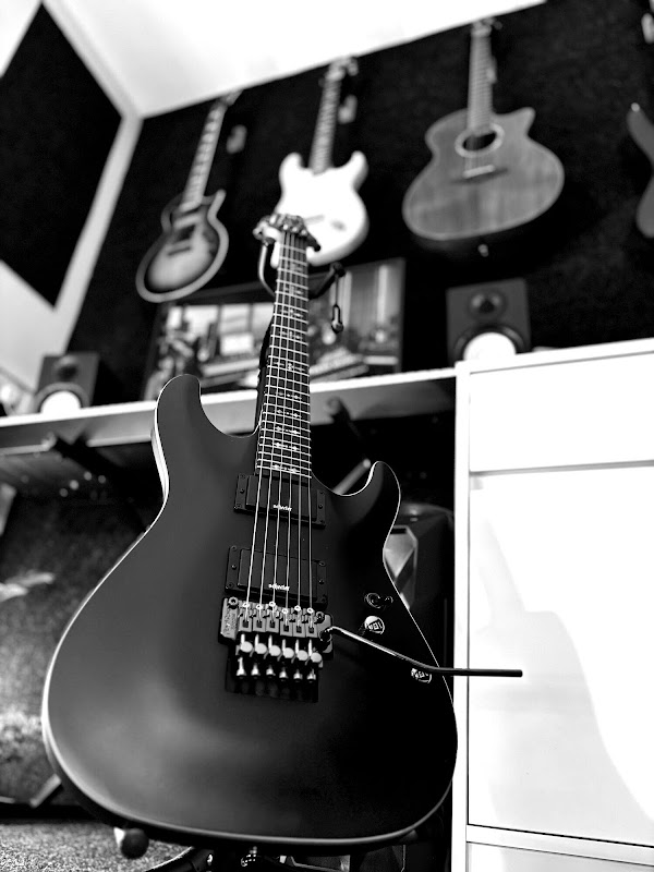 Black and white photo of a black electric guitar with a tremolo arm on a stand, with guitars hanging on the wall and studio speakers behind it