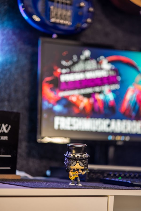 Small collectible figure of a top-hatted guitarist holding a gold guitar on the studio desk, in front of a screen showing the Fresh Music Aberdeen logo
