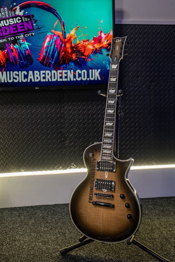 Sunburst electric guitar with Fresh Music Aberdeen printed on the body, on a stand beneath a wall-mounted screen showing the Fresh Music Aberdeen logo