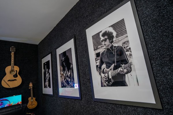 Framed black and white portraits of famous guitarists on the carpeted acoustic wall of the studio, with acoustic guitars hanging in the corner
