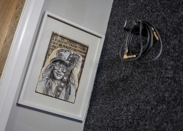 Framed hand-drawn sketch of a curly-haired guitarist in a top hat and sunglasses leaning against the wall, beside a coiled guitar cable on the carpet