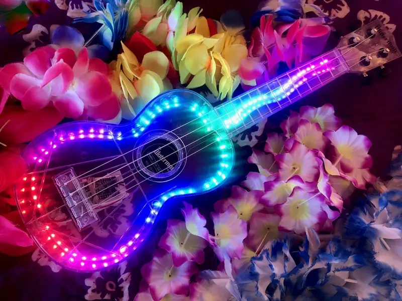 Clear ukulele lit up with rainbow LED lights, resting on a bed of colourful Hawaiian flower leis